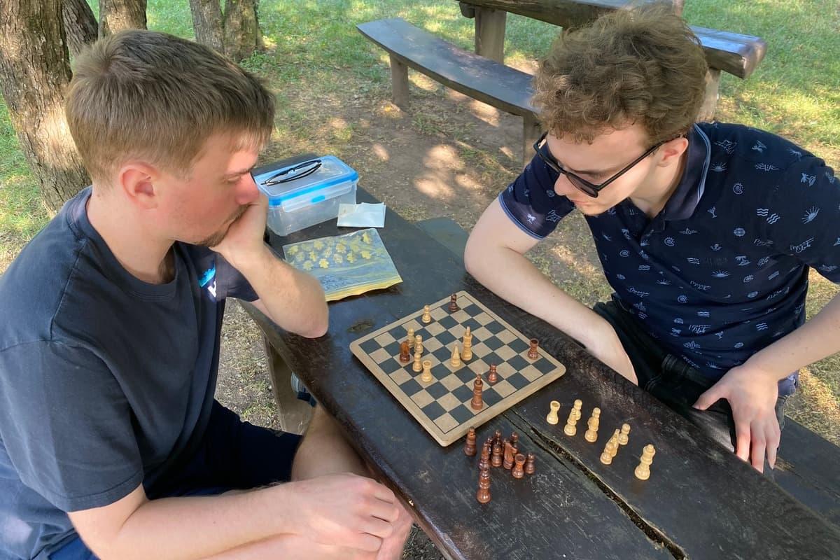 Two people playing chess at a park table stay sharp while the trees and benches behind them are blurred — original